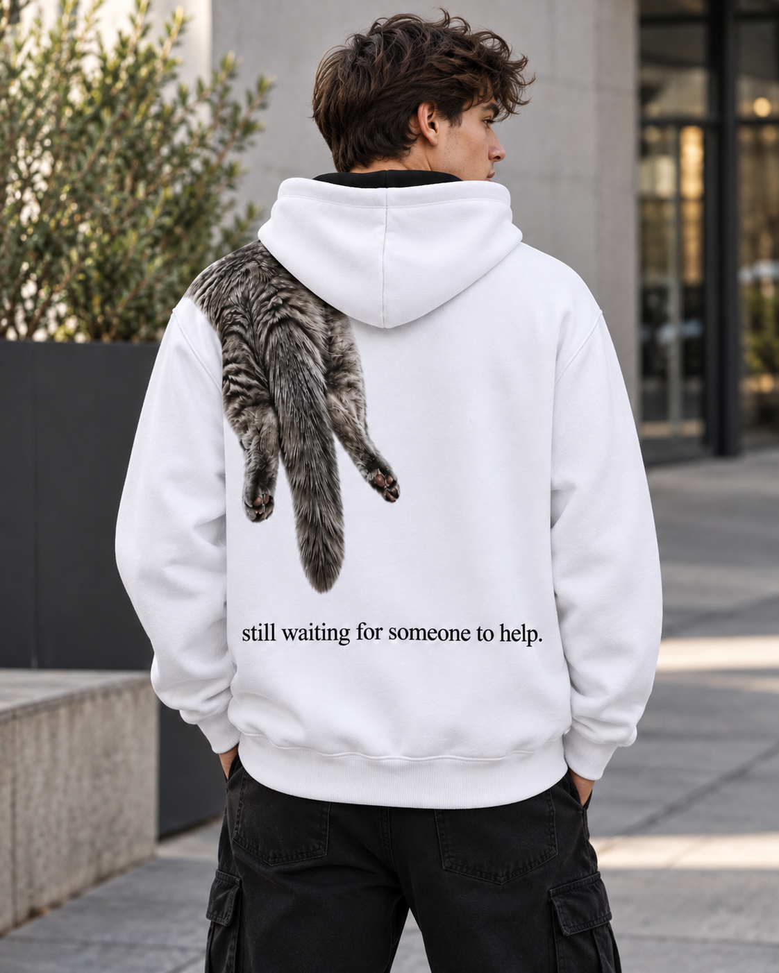 Back view of man wearing a white VZAEMOA graphic hoodie with grey shoulder cat artwork outdoors